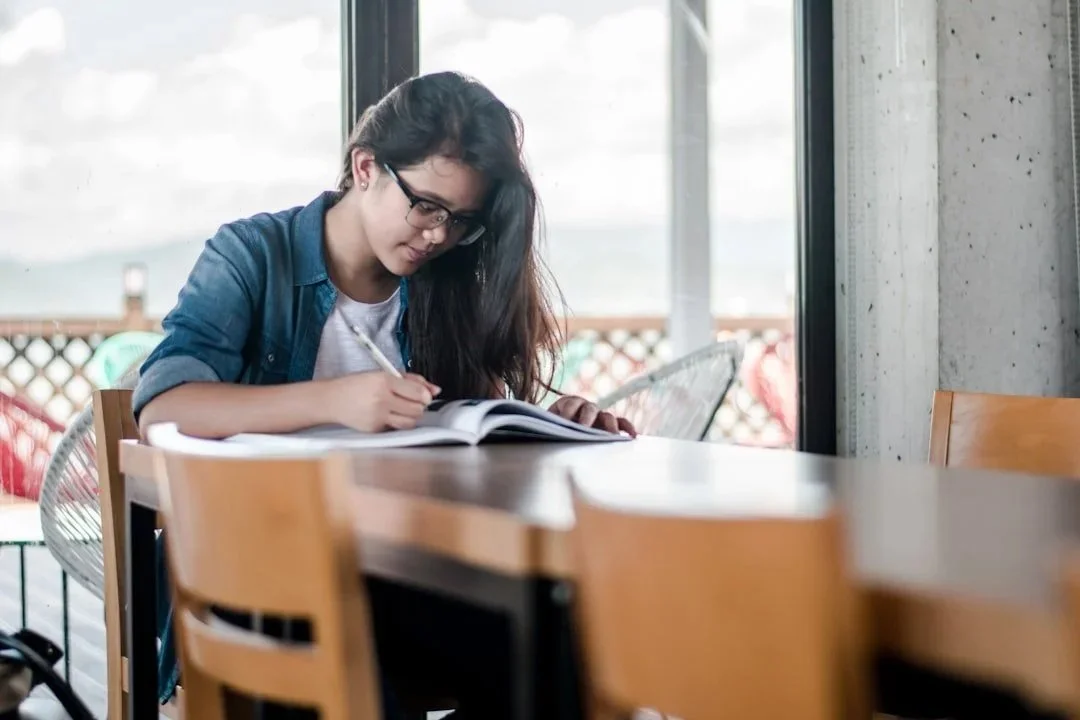 Student wearing glasses and writing in an open book at a table