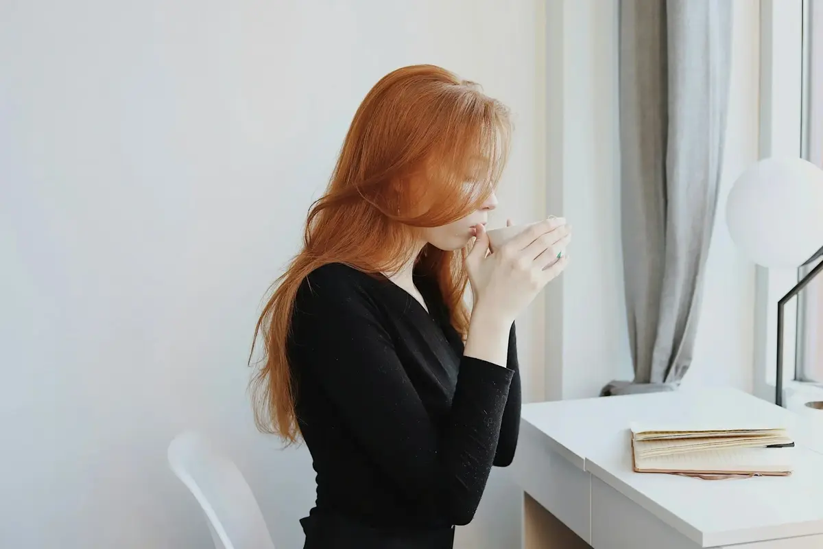 Red-haired person holding a mug beside a window