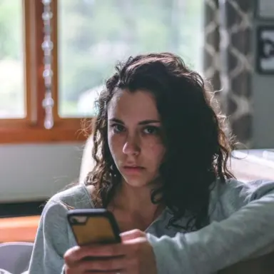Woman holding a phone while seated on a sofa beside a window