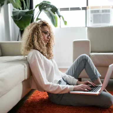 Woman using a laptop while sitting on the floor beside a sofa.
