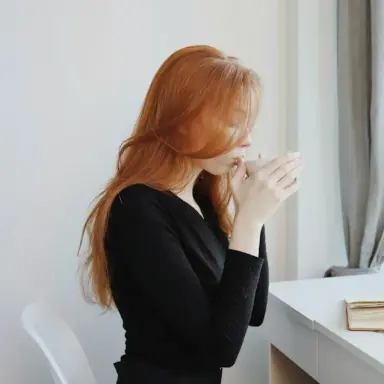 Woman seated by a window with her hands together near her face and a notebook on the desk.
