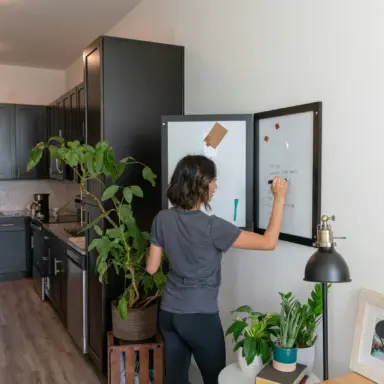 Person writing on a wall-mounted whiteboard beside a kitchen.