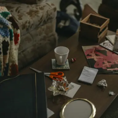 Coffee table covered with magazines, papers, scissors, and a mug.