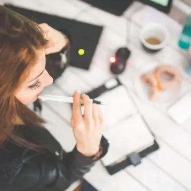 Woman holding a pen near her face while looking over papers on a desk.