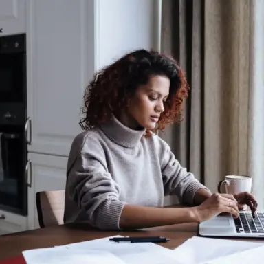 Woman working on a laptop at a table beside a window.